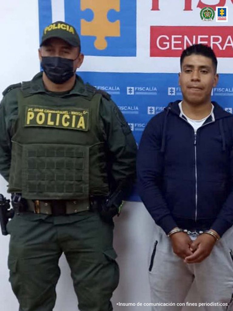 En la foto aparece de pie Luis Alberto Sarchi Carlosama, con los brazos adelante y esposado. Viste con buzo oscuro y pantalón de sudadera gris, zapatos tenis blancos con rojo y cordones oscuros. Al lado derecho de él hay un uniformado de la Policía Nacional y a su izquierda un servidor del CTI de la Fiscalía. Detrás de ellos hay un pendón institucional de la Fiscalía. 