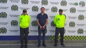 En la fotografía se aprecia de pie y con los brazos adelante y esposado a Luis Alfonso Montoya Molina. Viste camisa de manga corta negra, jean azul oscuro y zapatos negros. A lado y lado de él hay uniformados de la Policía Nacional. Detrás de ellos hay un pendón institucional de la Policía.