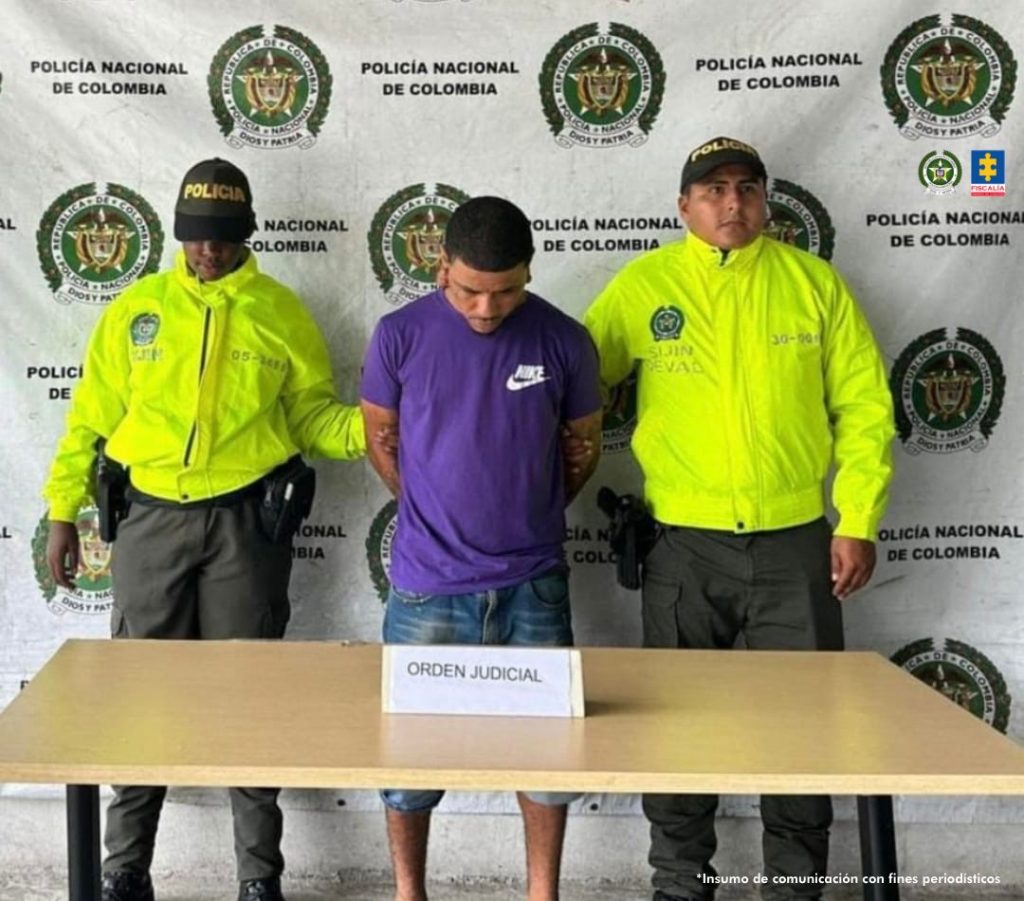 En la fotografía se aprecia de pie y con los brazos atrás a Luis Alfredo Orozco Carabalí, alias 3D. Viste camiseta de manga corta de color púrpura, con la palabra ‘Nike’ y el logo de esa marca en color blanco, en la parte superior izquierda del pecho; bermudas en jean azul desgastado. A sus extremos hay uniformados de la Policía Nacional. Delante de ellos hay una mesa y sobre ella hay un letrero que dice: ‘Orden judicial’. Detrás de ellos hay un pendón institucional de la Policía.