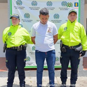 En la fotografía aparece una persona capturada, acompañado de un uniformado de la Policía Nacional y del Ejército Nacional.  En la parte posterior un banner con logos de la entidad.  