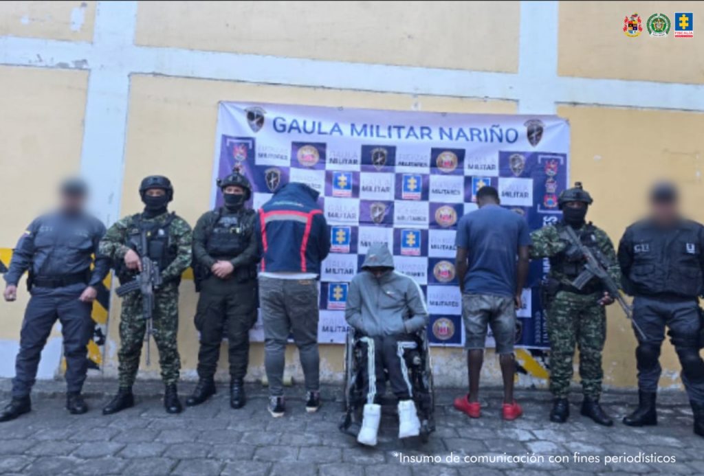 En la fotografía aparecen los presuntos implicados, dos de ellos de espaldas a cámara y uno de frente (el del centro) en silla de ruedas. Uno, viste chaqueta de color azul oscuro con franjas rojas verticales en la espalda; pantalón gris y zapatillas negras. Otro, en silla de ruedas, viste buso de color gris, con franjas blancas en las mangas, sudadera de color negro con franjas blancas a los lados y zapatillas blancas; y un tercero, viste con camiseta de manga corta, de color azul oscuro, bermudas de color gris y zapatillas rojas. Al extremo izquierdo de la fotografía hay un servidor del CTI de la Fiscalía y uniformados del Ejército y Policía Nacional. Al extremo derecho hay un servidor del CTI de la Fiscalía y un uniformado del Ejército. Detrás de ellos hay un pendón con logos de la Fiscalía y del Ejército.--
