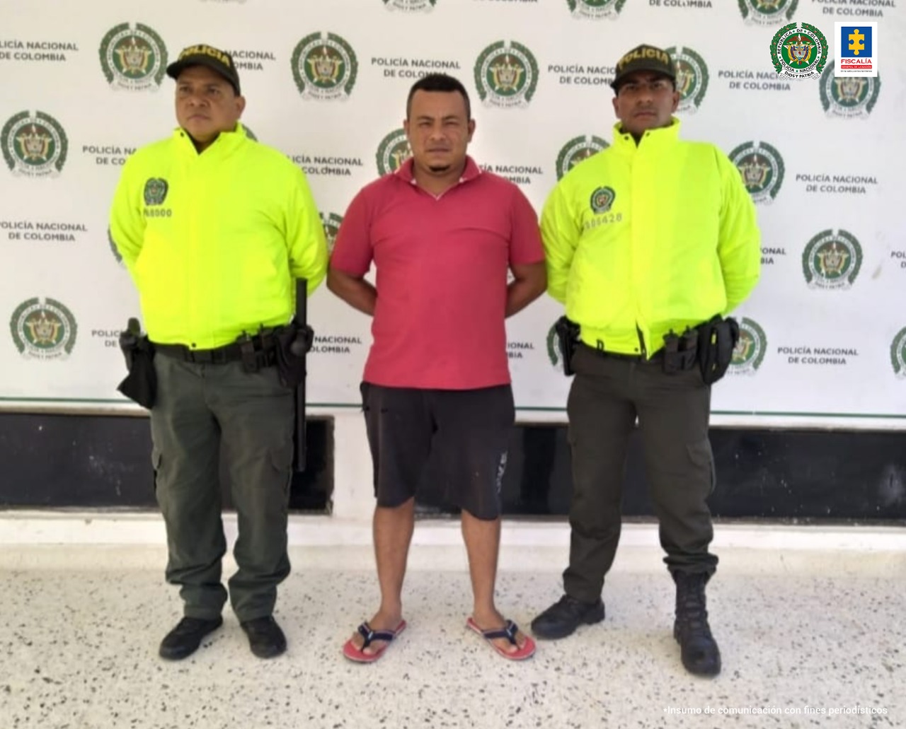 En la fotografía aparece un hombre capturado, acompañado de dos uniformados de la Policía Nacional. En la parte posterior un banner con logos de la entidad.