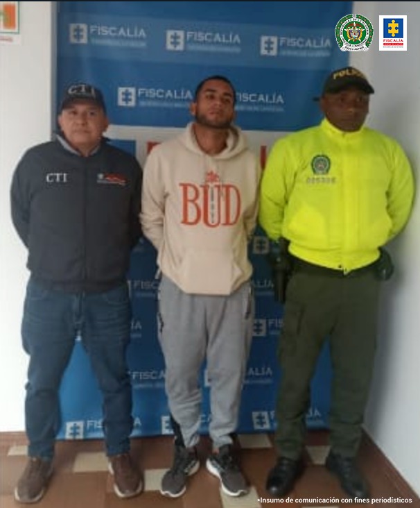 En la foto aparece de pie y con los brazos atrás Luis Fernando Gómez Palomino. Viste buso claro con estampado rojo a la altura del pecho; pantalón de judadera gris, tenis negros con blanco. Al lado derecho de él hay un servidor del CTI de la Fiscalía y a su izquierda un uniformado de la Policía Nacional. Detrás de ellos hay un pendón interinstitucional de la Fiscalía.