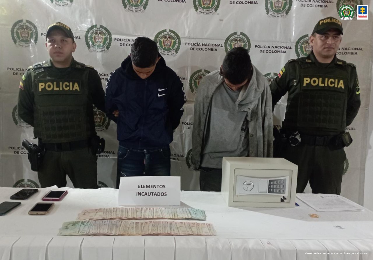 En la foto aparecen Luis Fernando Murillo Marín y Luis Alberto Tulande Ramírez acompañados de dos policías a los extremos y de fondo los logos de la Policía Nacional. En una mesa se puede ver dinero en efectivo y una caja fuerte y cuatro celulares.