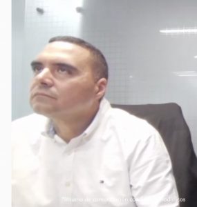 Se ve al hombre de medio cuerpo, sentado, mirando hacia arriba. Al fondo se ve un panel de vidrio. 