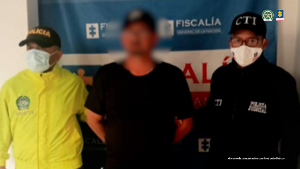 En la fotografía se aprecia de pie y con los brazos atrás a Luis Germán Velasco Ángel. Viste con camiseta, pantalón y gorra de color negro; así como zapatillas blancas. Al lado izquierdo de él hay un servidor del CTI de la Fiscalía. Al lado derecho un uniformado de la Policía Nacional. Detrás de ellos hay un pendón institucional de la Fiscalía.