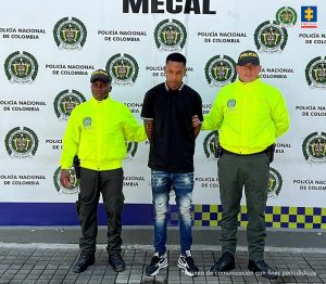 En la fotografía se aprecia de pie y con los brazos atrás a Luis Mateo Mina Córdoba. Viste camisa negra con cuello y manga corta. Jean azul desgastado y zapatillas negras con blanco. A sus extremos hay uniformados de la Policía Nacional. Detrás de ellos hay un pendón institucional de la Policía.