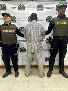 Se observa al capturado de espaldas y a cada costado un funcionario de Policía Nacional.  Atrás un pendón de Policía nacional