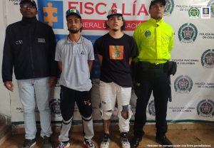 Se observan los dos detenidos en fila y a un lado de ellos un funcionario del CTI y al otro, uno de Policía nacional.  Atras un pendón de Fiscalía y uno de Policía Nacional.