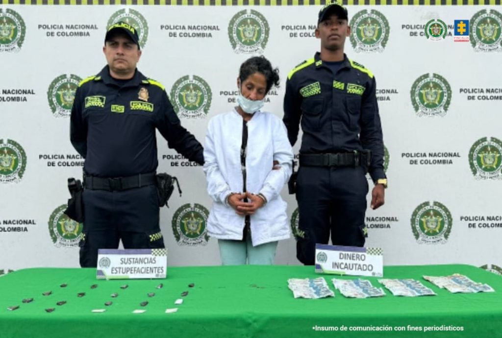 En la fotografía se aprecia de pie, con los brazos adelante y esposada a Magda Lorena Morales. Viste chaqueta blanca y pantalón verde. Usa tapabocas azul. A sus extremos hay uniformados de la Policía Nacional. Delante de ellos hay una superficie con mantel verde y sobre ella hay bolsas plásticas pequeñas y dinero en efectivo. Asimismo, hay dos letreros que dicen: ‘Sustancias estupefacientes’ y ‘Dinero incautado’. Detrás de ellos hay un pendón institucional de la Policía.