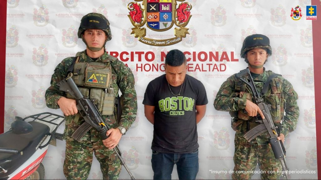 El imputado se encuentra en custodia por dos uniformados del Ejército Nacional. Detrás hay un backing del Ejército.