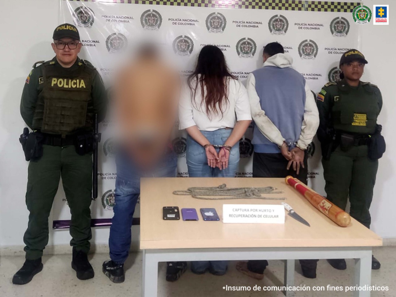 Se observan tres capturados en fila y de espaldas. Dos hombres y una mujer. En cada extremo de la fila un funcionario de Policía Nacional. Al frente una mesa con un cartel que dice: captura por hurto - recuperación de celular. En la misma reposan varios elementos incautados. Al fondo un backing de Policía Nacional