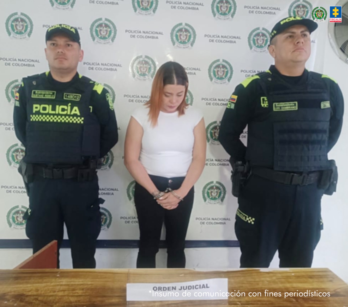 En la fotografía se aprecia de pie, con los brazos adelante y esposada a María Corona Arango Grajales. Viste con blusa blanca de manga en el hombre y pantalón negro. Usa cabello de color naranja y raiz oscura. A sus extremos hay uniformados de la Policía Nacional. Delante de ellos hay un superficie de color madera y sobre ella un letrero que dice: ‘Orden judicial’. Detrás de ellos hay un pendón institucional de la Policía.