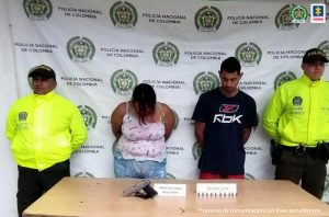 En la foto aparecen, con los brazos atrás, María Isabel Henao Betancourth y Héctor Fabio Alfaro Navarro. La mujer tiene la cabeza agachada; viste una blusa rosada, de tiras, con estampado y pantalón jean azul claro. El hombre tiene la mirada hacia el piso. Viste camiseta de manga corta negra con estampado de letras blancas; se aprecia pantalón rojo. A lado y lado de ellos hay uniformados de la Policía Nacional. Frente a ellos hay una mesa y sobre ella hay una pistola y varios cartuchos; así como dos letreros que dicen ‘Munición’ y ‘arma de fuego incautada’. Detrás de ellos hay un pendón institucional de la Policía Nacional.