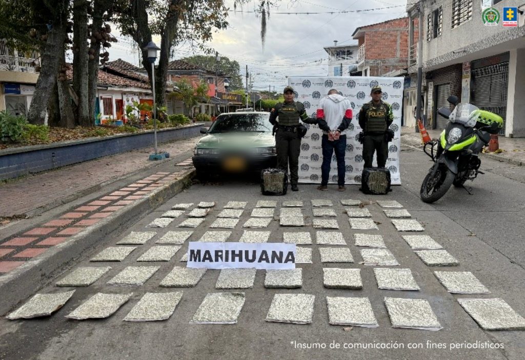 La fotografía es tomada en exteriores y se aprecia de espaldas a la cámara, con los brazos atrás y esposado a Mario Alexander López Mesías. Viste con buso de color blanco y mangas largas con los colores rojo y negro; pantalón negro. A sus extremos hay uniformados de la Policía Nacional. A los pies de los uniformados y sobre el suelo hay dos paquetes envueltos en plástico negó. Asimismo, sobre el suelo hay varios paquetes de plástico, rectangulares y delgados con contenido vegetal. Sobre dichos paquetes hay un letrero que dice: ‘ Marihuana’. En la parte posterior hay un vehículo particular, un pendón institucional de la Policía y al lado una motocicleta de la misma institución.