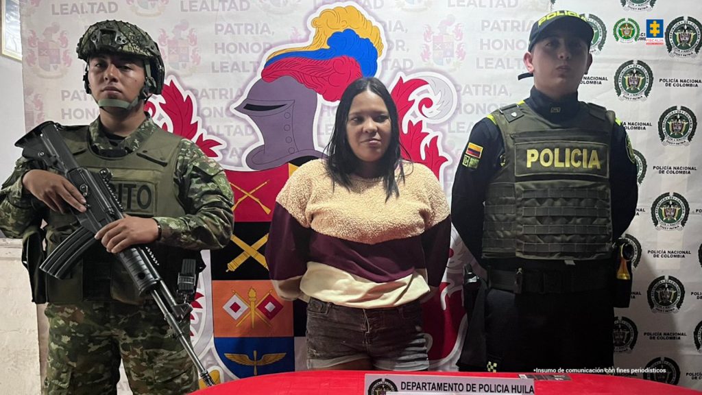 En la fotografía aparece una mujer capturada, acompañada de dos uniformados de la Policía.  En la parte posterior un banner con logos de la entidad.    