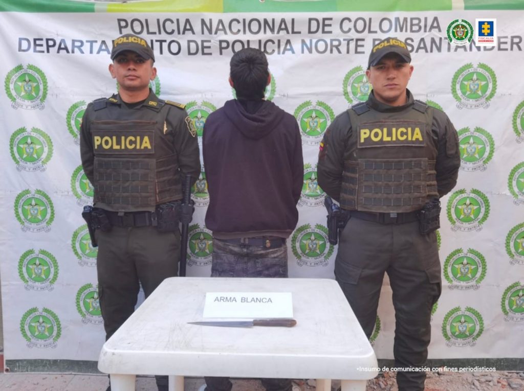 En la imagen se observa al capturado junto a dos uniformados de la Policía Nacional. Frente a ellos una mesa donde se encuentra el arma cortopunzante utilizada para la acción ilícita. En la parte posterior se observa el banner que identifica