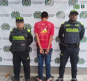 capturado de espaldas custodiado por dos policías. Detrás banner de Policía Nacional.