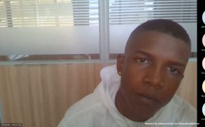  En la fotografía está el rostro de un hombre joven, moreno, de cabellos cortos crespo y negro, vestido con un buzo blanco, una cadena dorada y un arete dorado. 