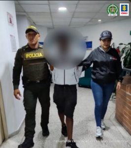 En la fotografía se aprecia en un pasillo, en acción de caminata y con los brazos atrás, al presunto implicado. Viste buso blanco con cremallera negra, bermuda negra y zapatillas negras con suela blanca. Al lado izquierdo de él hay una servidora del CTI de la Fiscalía y al lado derecho un uniformado de la Policía Nacional.