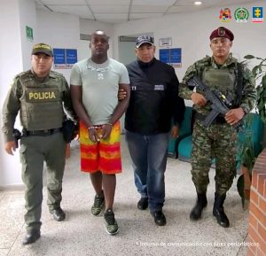 En la fotografía se aprecia en acción de caminata por un pasillo, con los brazos adelante y esposado a Jefferson Herrera Valencia. Viste camisa de manga corta azul claro, bermuda de color rojo y amarillo y zapatillas negras con zuela blanca. Al lado izquierdo de él hay un servidor del CTI de la Fiscalía y un uniformado del Ejército Nacional. Al lado derecho hay un uniformado de la Policía Nacional.