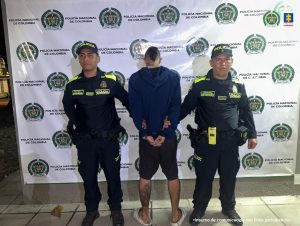 Texto Alternativo: capturado de espaldas custodiado por dos policías. Detrás banner de Policía Nacional.