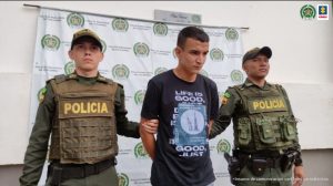 en la imagen se ve una persona detenida bajo custodia de dos integrantes de la Policía Nacional. Detrás suyo un backing institucional.