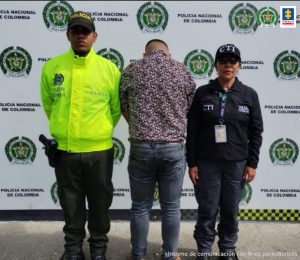 En la fotografía se aprecia de pie y de espaldas a la cámara a Mauricio Alejandro Baena Saavedra. Viste camisa estampada, jean gris y zapatos color miel. Al lado izquierdo de él hay una servidora del CTI de la Fiscalía y al lado derecho de él hay un uniformado de la Policía Nacional. En la parte posterior de ellos hay un pendón institucional de la Policía.