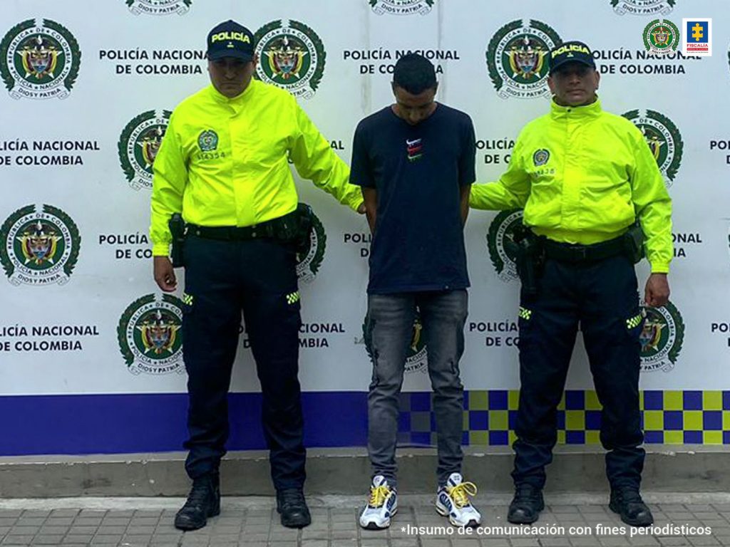 En la fotografía aparece de pie, con los brazos atrás y la cabeza agachada Mauricio Reyes Cobo. Camiseta de manga corta negra, jean gris desgastado y zapatillas blancas con negro y cordones amarillos. A lado y lado de él hay uniformados de la Policía Nacional. Detrás de ellos hay un pendón institucional de la Policía Nacional.