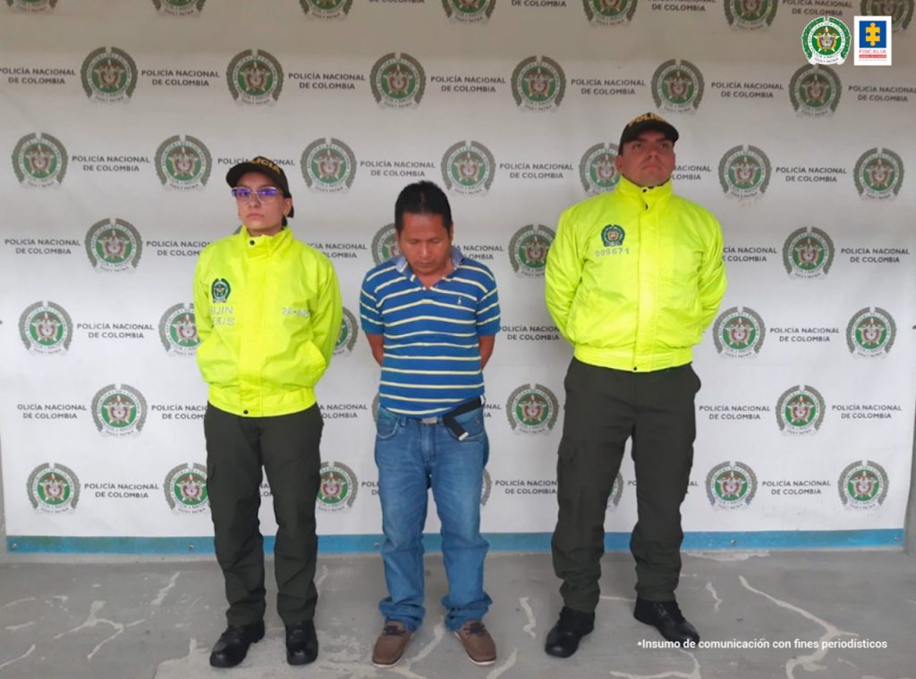 Se observa un hombre vestido con camiseta de rayas horizontales en azul y blanco, de frente y a cada costado un funcionario de Policía Nacional. Al fondo un pendón de Policía Nacional