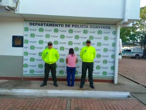 capturada de espaldas custodiada por dos policías. Detrás banner de la Policía Nacional.