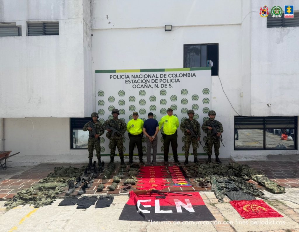 Se visualiza al capturado junto a uniformados de la Policía y Ejército Nacional. Frente a ellos el material incautado (uniformes, brazaletes, panfletos y banderas del ELN)