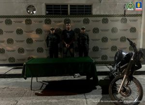 En la fotografía se aprecia de pie, con los brazos adelante y esposado a Michael Steven Castañeda Ruiz. Viste camiseta negra de manga corta, con estampado a la altura del pecho; jean azul desgastado en los muslos. A sus extremos hay uniformados de la Policía Nacional. Delante de ellos hay una mesa con mantel verde vacía. Al lado de ella hay una motocicleta. Detrás de ellos hay un pendón institucional de la Policía.