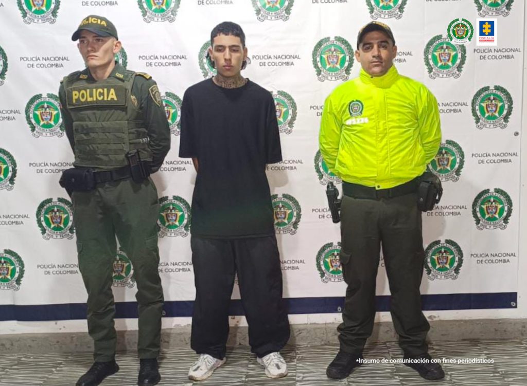 En la fotografía se aprecia a Miguel Ángel Barona Orozco de pie, con los brazos atrás. Viste camiseta de manga corta y pantalón negro; zapatillas blancas. A lado y lado de él hay uniformados de la Policía Nacional. Detrás de ellos hay un pendón institucional de la Policía.