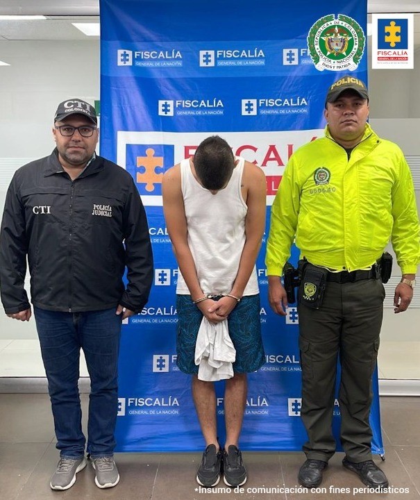 En la foto se ve al capturado con la cabeza agachada, esposado en pantaloneta y camiseta sin mangas y una prenda en la mano, custodiado a lado y lado por un Policía y un investigador del CTI de la Fiscalía uniformados, detrás un banner de la Fiscalía.