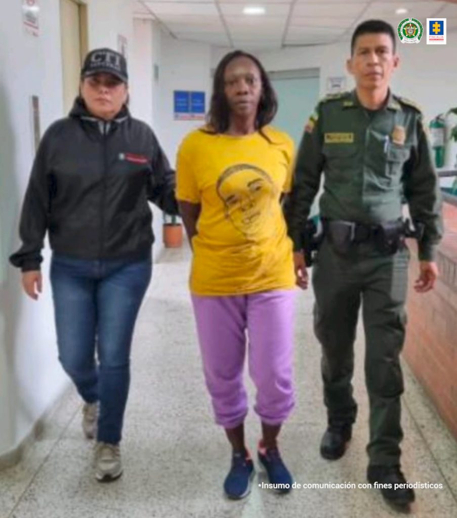 En la fotografía se aprecia a Mileydi Vallecilla Hinestroza en acción de caminata, por un pasillo, con los brazos atrás. Viste camiseta de manga corta color mostaza con estampado del rostro de un hombre en color negro; pantalón de sudadera color violeta y tenis oscuros. Al lado derecho de ella hay un servidor del CTI de la Fiscalía y a la izquierda un uniformado de la Policía.