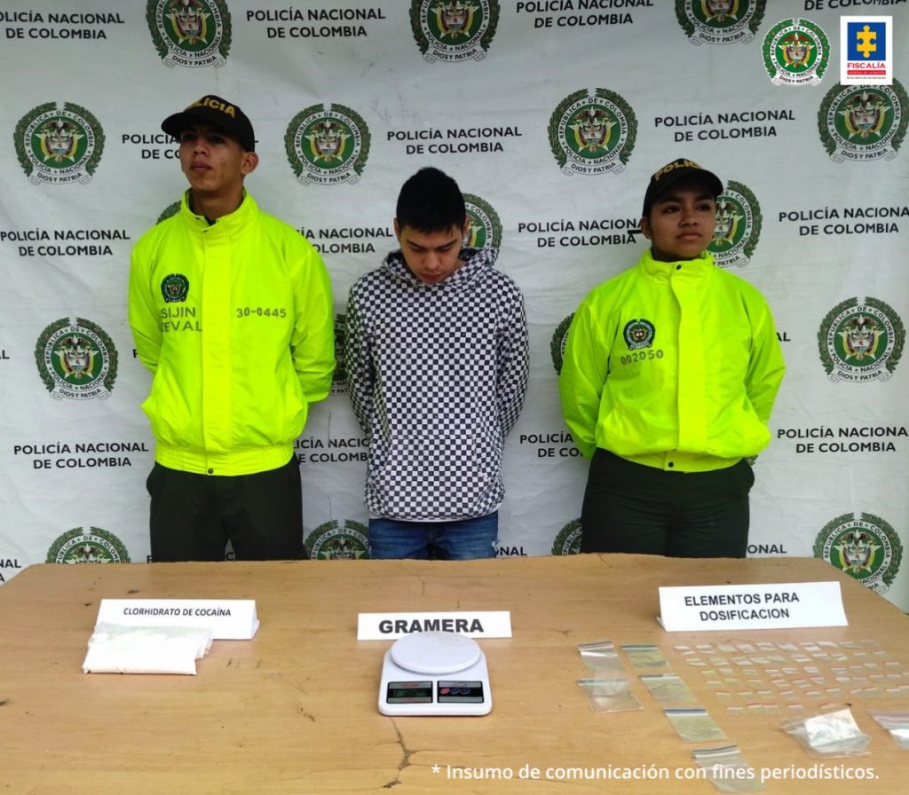 En la foto aparece el hoy imputado con los brazos atrás, la cabeza agachada; vestido con un camibuso estampado con cuadros grises y negros. A lado izquierdo y derecho aparecen dos integrantes de la Policía Nacional. Delante de ellos hay una mesa y sobre ella hay una gramera, elementos para dosificación y clorhidrato de cocaína. Detrás de ellos hay un pendón institucional de la Policía Nacional.