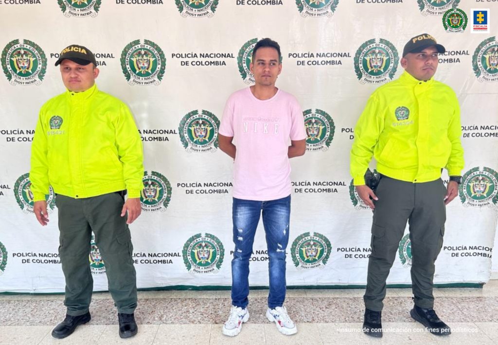 En la fotografía aparece un capturado junto a personal de Fiscalía General de la Nación y Policía Nacional. En la parte posterior de la imagen se ven banners de Policía Nacional y la Fiscalía General de la Nación.