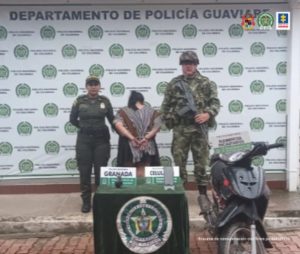 capturada de espaldas esposada y custodiada por un soldado y un policía, delante una mesa con material incautado y detrás banner de la Policía Nacional.