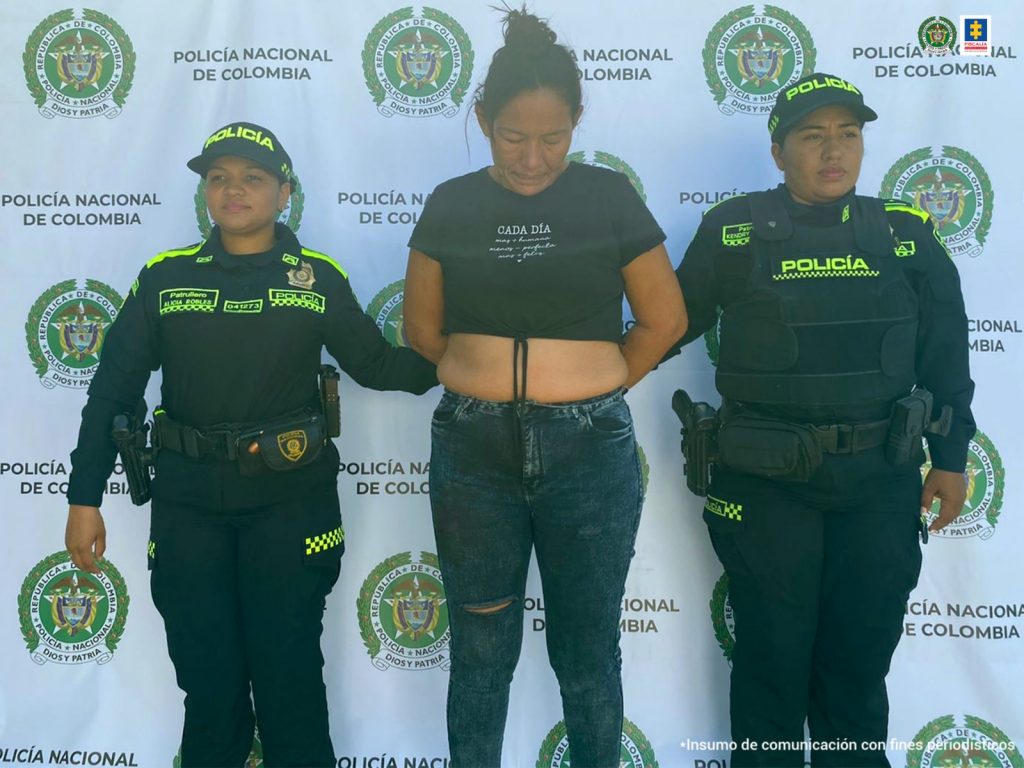 La procesada tiene una blusa de color negro, está esposada y acompañada por dos funcionarios de la Policía. De fondo está el pendón institucional de la Policía.