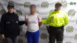 En la fotografía aparece de pie y los brazos atrás la abuela investigada. Viste blusa rosada de manga corta y pantalón azul. Al lado derecho hay una servidora del CTI de la Fiscalía. Al lado izquierdo de ella hay una uniformada de la Policía Nacional. Detrás de ellos hay un pendón institucional de la Policía.