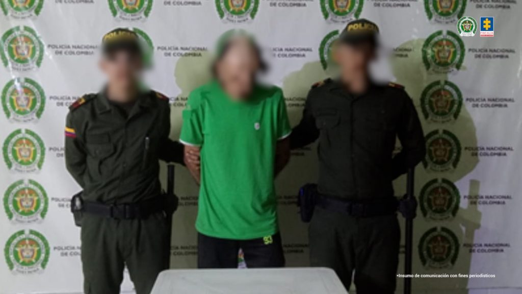 En la fotografía se aprecia de pie y con los brazos atrás al imputado. Viste camiseta de manga corta y de color verde; pantalón orcuro. A sus extremos hay uniformados de la Policía Nacional. Delante de ellos hay una superficie clara. Detrás de ellos hay un pendón institucional de la Policía.