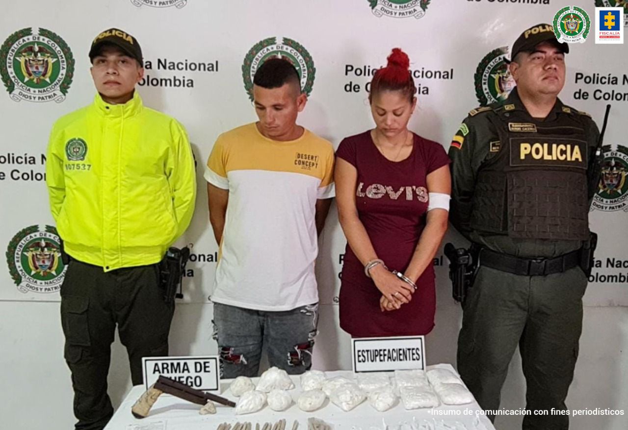 En la imagen están los dos detenidos con sus manos en la espalda, esposados, acompañados de dos uniformados de la Policía Nacional. Hay una mesa donde está el estupefaciente que les fue hallado en el momento de su captura y un arma de fuego. 