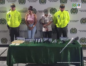 Fiscal Martha Mancera, Martha Mancera, Fiscal Mancera En la fotografía se aprecia de pie a Marcela González Martínez y Néstor Enrique Gómez Tovar. La mujer viste con blusa de manga siza rosada y jean azul claro ajustado. El hombre viste camisa de manga corta blanca con rayas horizontales de colores y pantalón oscuro. A lado y lado de ellos hay uniformados de la Policía Nacional. Delante de ellos hay una mesa con mantel verde y sobre ella se aprecian cuatro armas de fuego, tres proveedores, munición y unas placas. Asimismo, varios letreros que dicen: ‘Mercancía incautada’, ‘Munición’, ‘Armas de fuego’. Detrás de ellos hay un pendón institucional de la Policía.