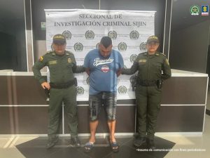 Se visualiza al capturado junto a dos uniformados de la Policía Nacional: Detrás de ellos el banner que identifica a la Policía Nacional.
