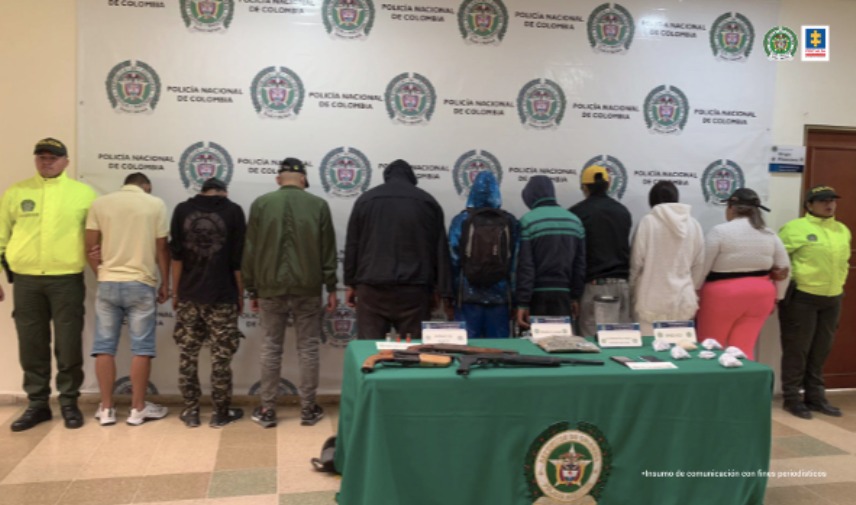 Se observa una fila de nueve personas, todos de espaldas, dentro de las cuales los presuntos implicados. A lado y lado de la fila un funcionario de Policía Nacional. Al fondo un pendón de Policía Nacional.