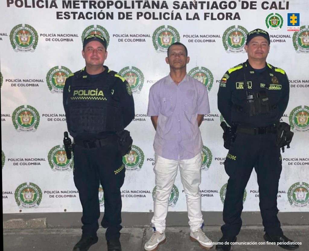 capturado de frente custodiado por dos policías. Detrás banner de la Policía Nacional.