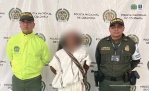 En la fotografía aparece un hombre capturado, acompañado de dos uniformados de la Policía Nacional. En la parte posterior un banner con logo de la entidad.