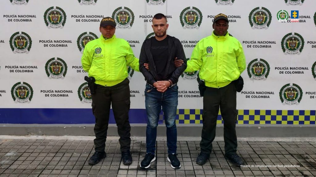 En la fotografía se aprecia de pie, con los brazos adelante y esposado a Néstor Fabio Vásquez Mejía. Viste camiseta y buso de manga larga y de color negro; jean azul y zapatillas negras con zuela blanca. A sus extremos hay uniformados de la Policía Nacional. Detrás de ellos hay un pendón institucional de la Policía.