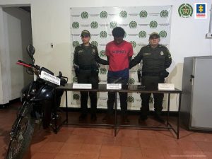En la foto aparece de pie, con los brazos atrás y la cabeza agachada, Néstor José Silva Herrera. Esta persona viste camiseta roja de manga corta y pantalón oscuro. A lado y lado de él hay uniformados de la Policía Nacional. Delante de ellos hay una mesa con 3 avisos que dicen: ‘celular recuperado’, ‘Arma’ y ‘hurto calificado’. Al lado derecho de la mesa hay una motocicleta con un letrero que dice: ‘Motocicleta recuperada’. Detrás de ellos hay un pendón institucional de la Policía.
